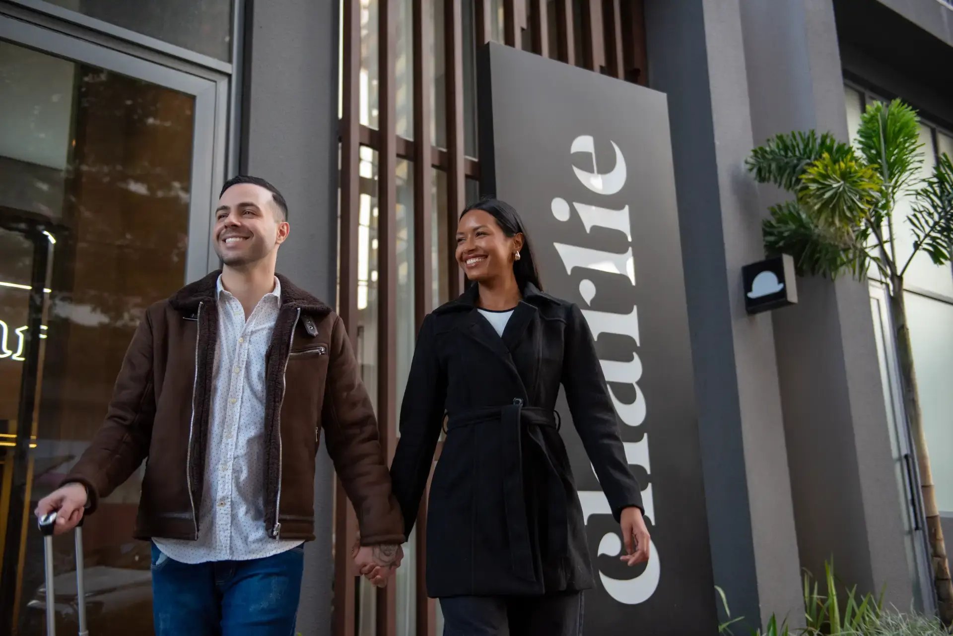 Casal sorridente caminhando de mãos dadas em frente a uma fachada moderna com plantas e porta de vidro, em ambiente externo com luz natural.