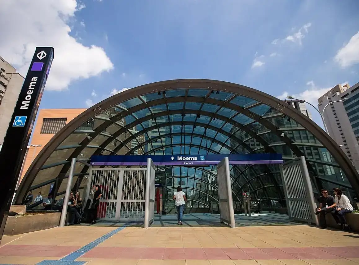 Entrada da estação de metrô Moema em São Paulo, com estrutura de vidro e pessoas ao redor, sob céu azul com algumas nuvens.