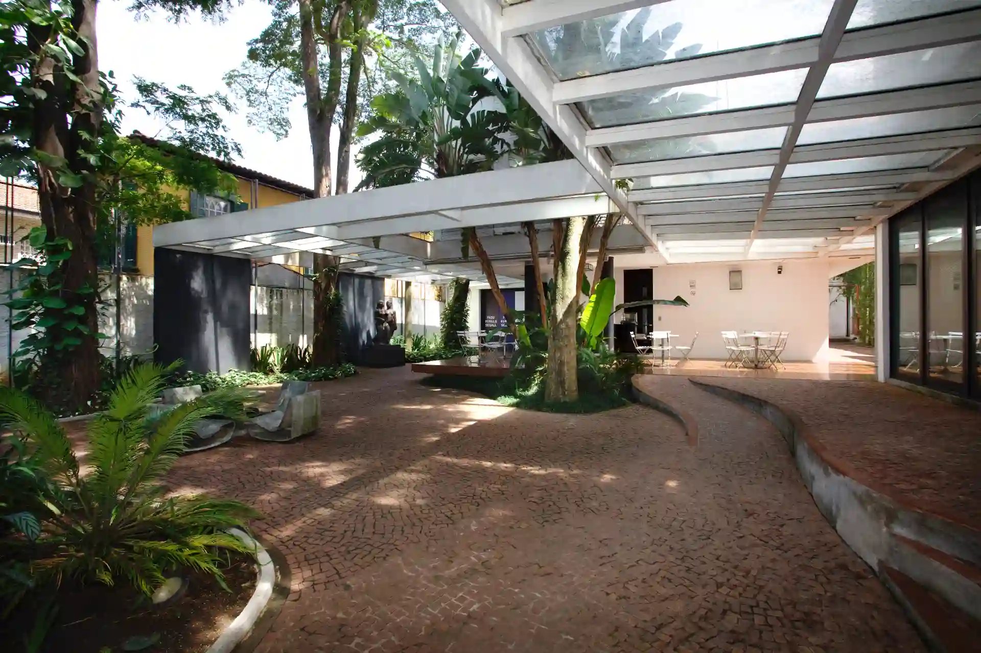 Áreas externas de um condomínio com espaço de convivência ao ar livre, vegetação exuberante, árvores, plantas decorativas, estruturas modernas de vidro e vidro, piso de pedra, ambiente acolhedor e bem cuidado.