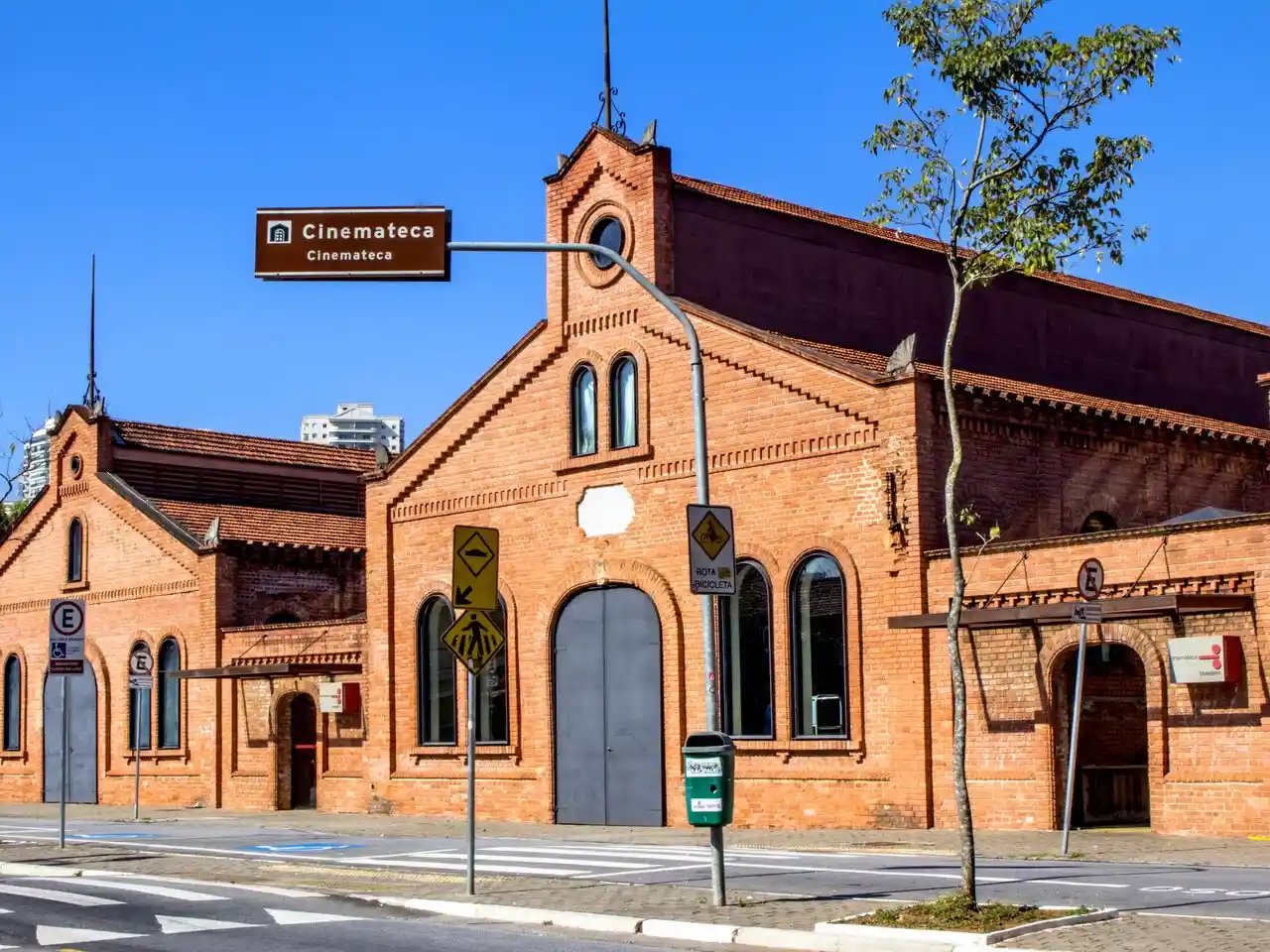 Imagem da fachada de um antigo prédio vermelho com arquitetura em estilo gótico, localizado próximo a uma rua com sinalização urbana, árvores e céu claro ao fundo, Representando uma cinematográfica arquitetura urbana no Brasil.