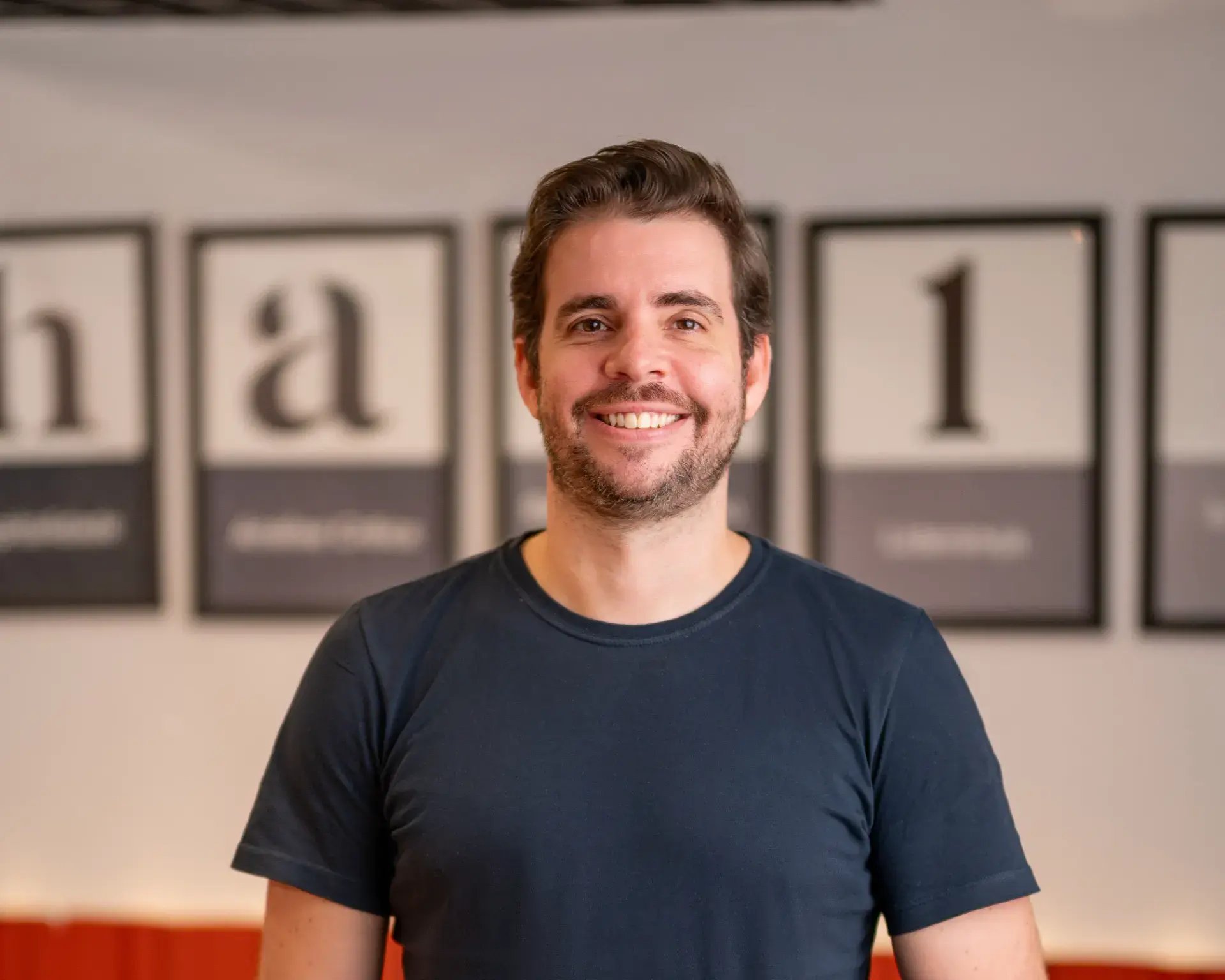 Homem sorridente com camiseta azul em ambiente de escritório, ao fundo há quadros com numeração e textos desfocados na parede.