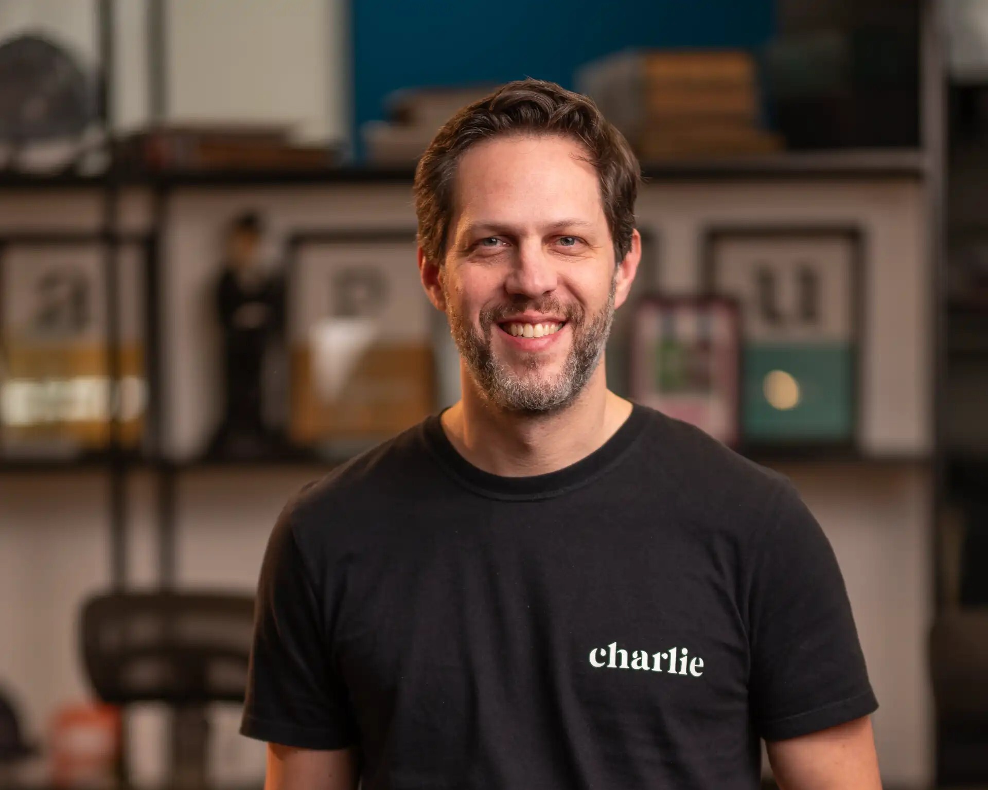 Homem sorrindo sentado ao lado de uma mesa de madeira, com camiseta preta escrita “charlie” e boné apoiado ao lado em um ambiente de escritório.