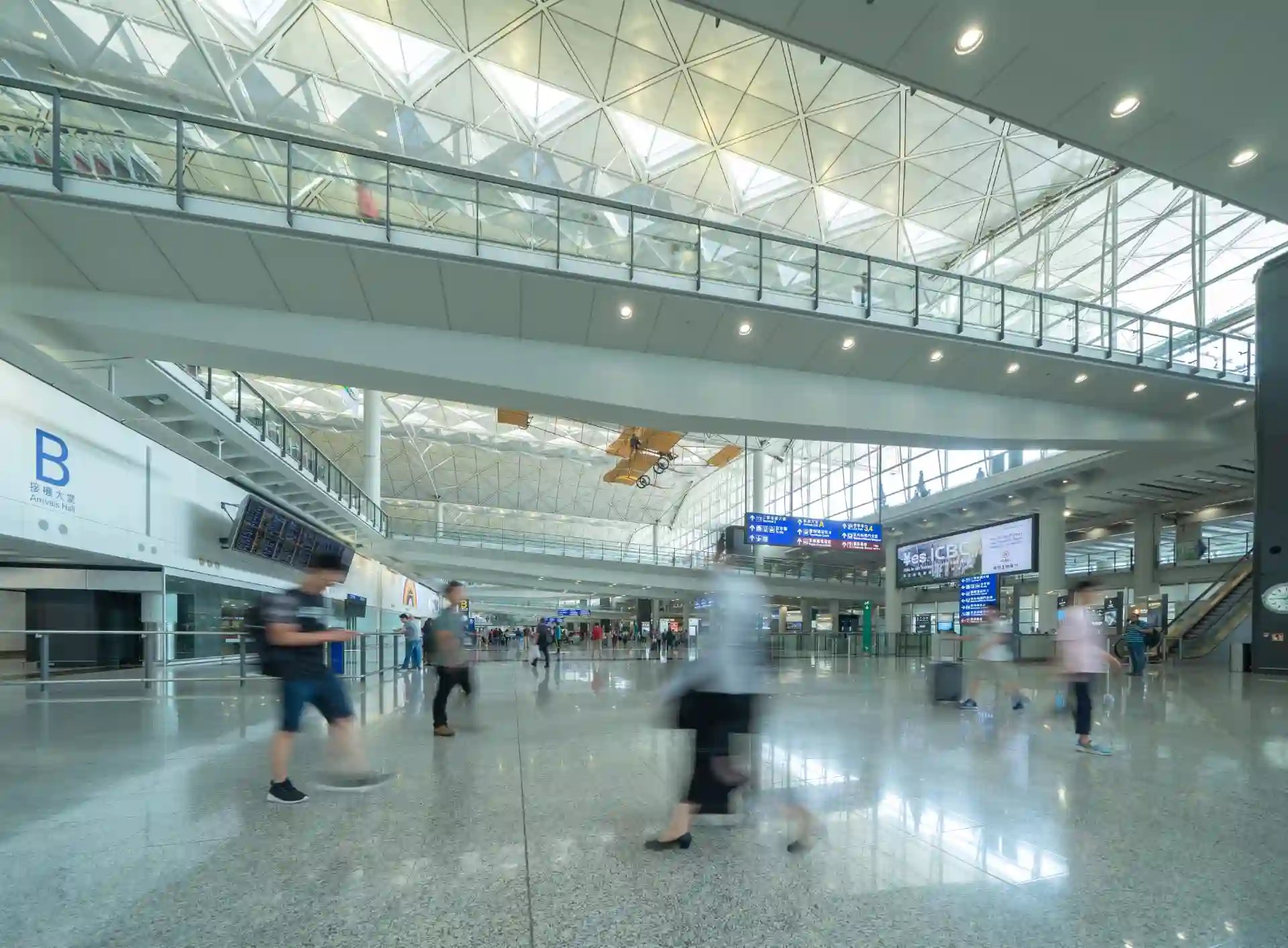 Vista ampla do interior de um aeroporto movimentado com várias pessoas caminhando, um avião decolando no teto e painéis de informações digitais.