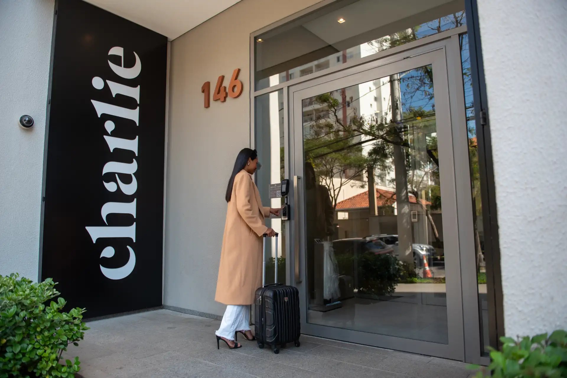 Mulher em frente a uma porta de hotel com a placa “Clara Hotel” e número 146, usando um telefone e ao lado de uma mala de viagem preta no saguão externo.