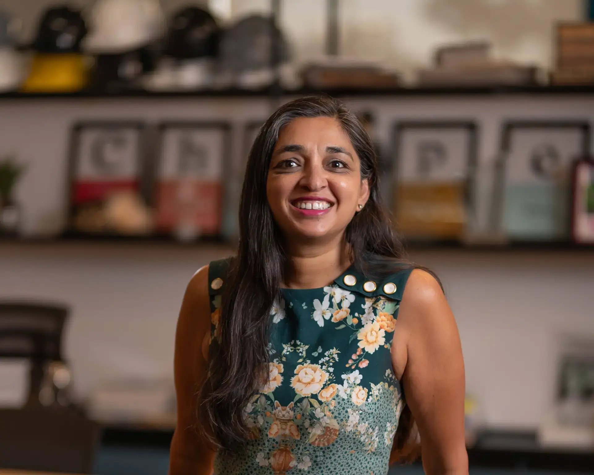 Mulher sorrindo em ambiente de escritório com prateleiras ao fundo, vestindo vestido floral verde sem manga e brincos discretos. Foto corporativa