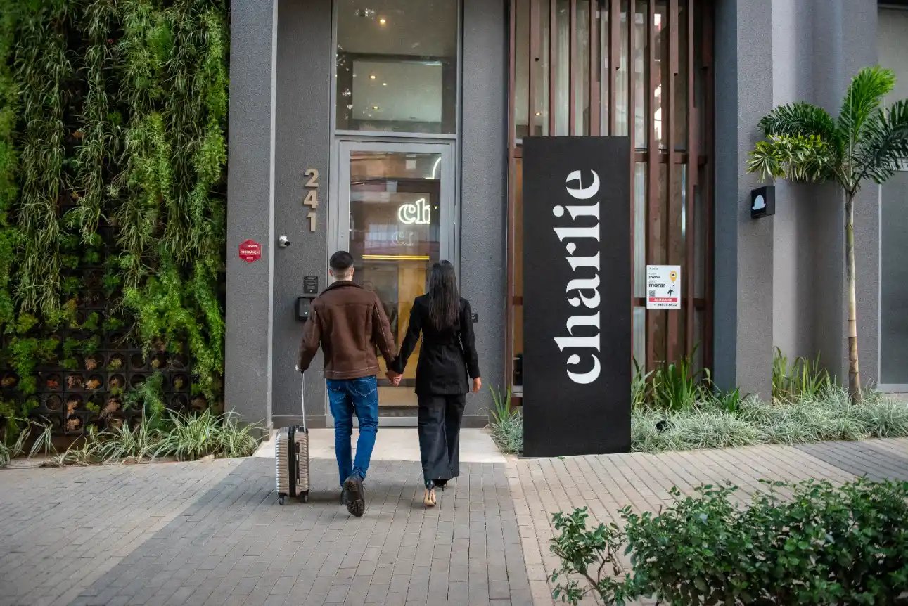 Pessoas caminhando em frente a uma entrada de hotel com placa “Charlie” e número 2411, cercada por plantas e jardim ao lado da fachada.