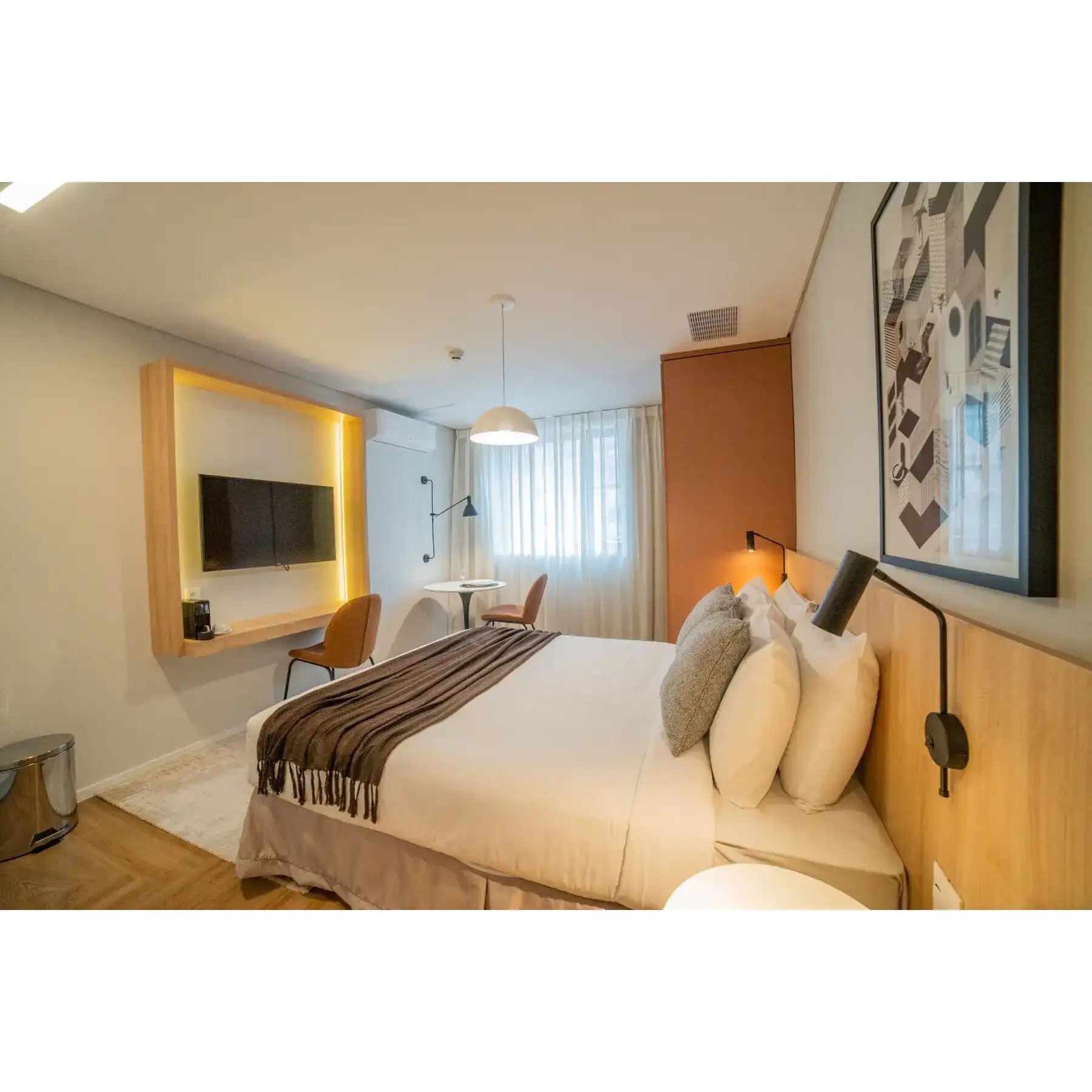Imagem de um quarto de hotel bem decorado com cama de casal, televisão na parede com iluminação de LED, mesa de canto, cadeira e decoração moderna.