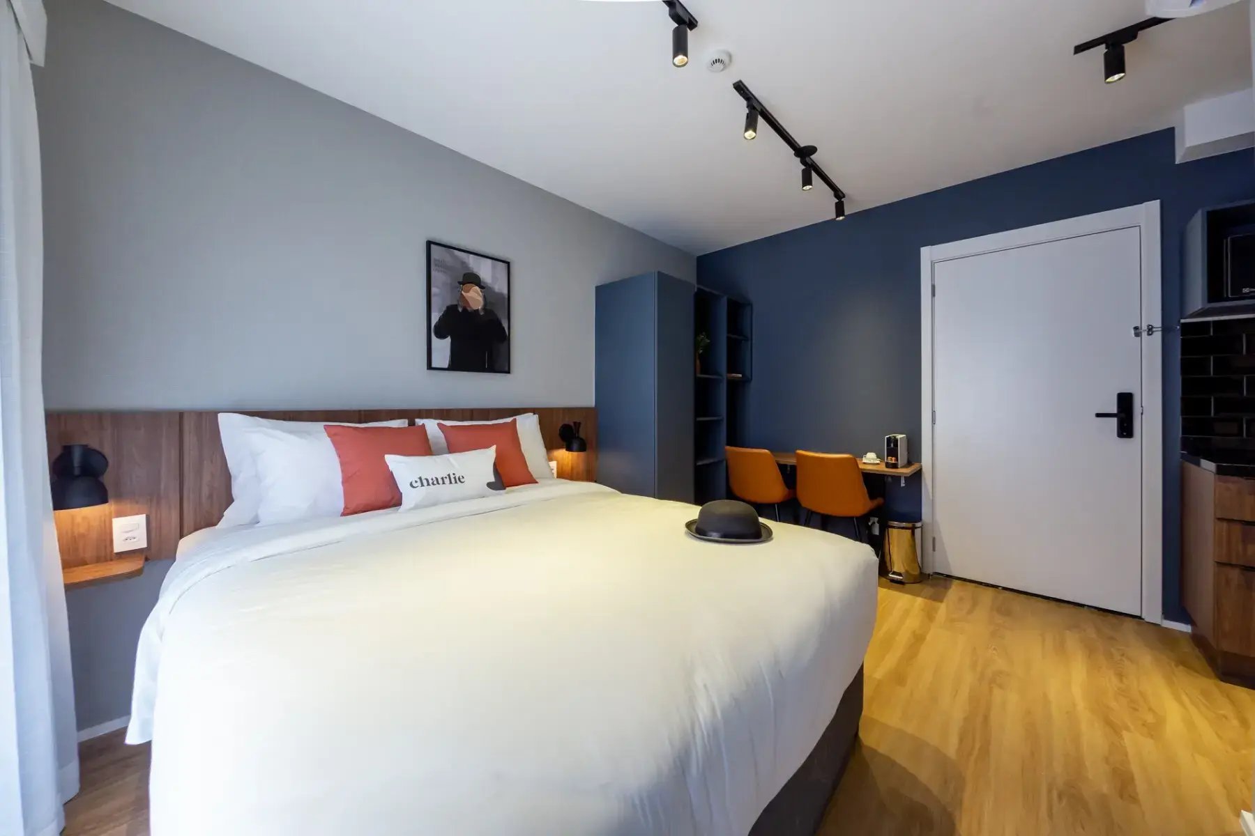 Quarto de hotel moderno com cama de casal, decoração azul, escrivaninha com cadeira amarela e uma parede com quadro de fotografia.