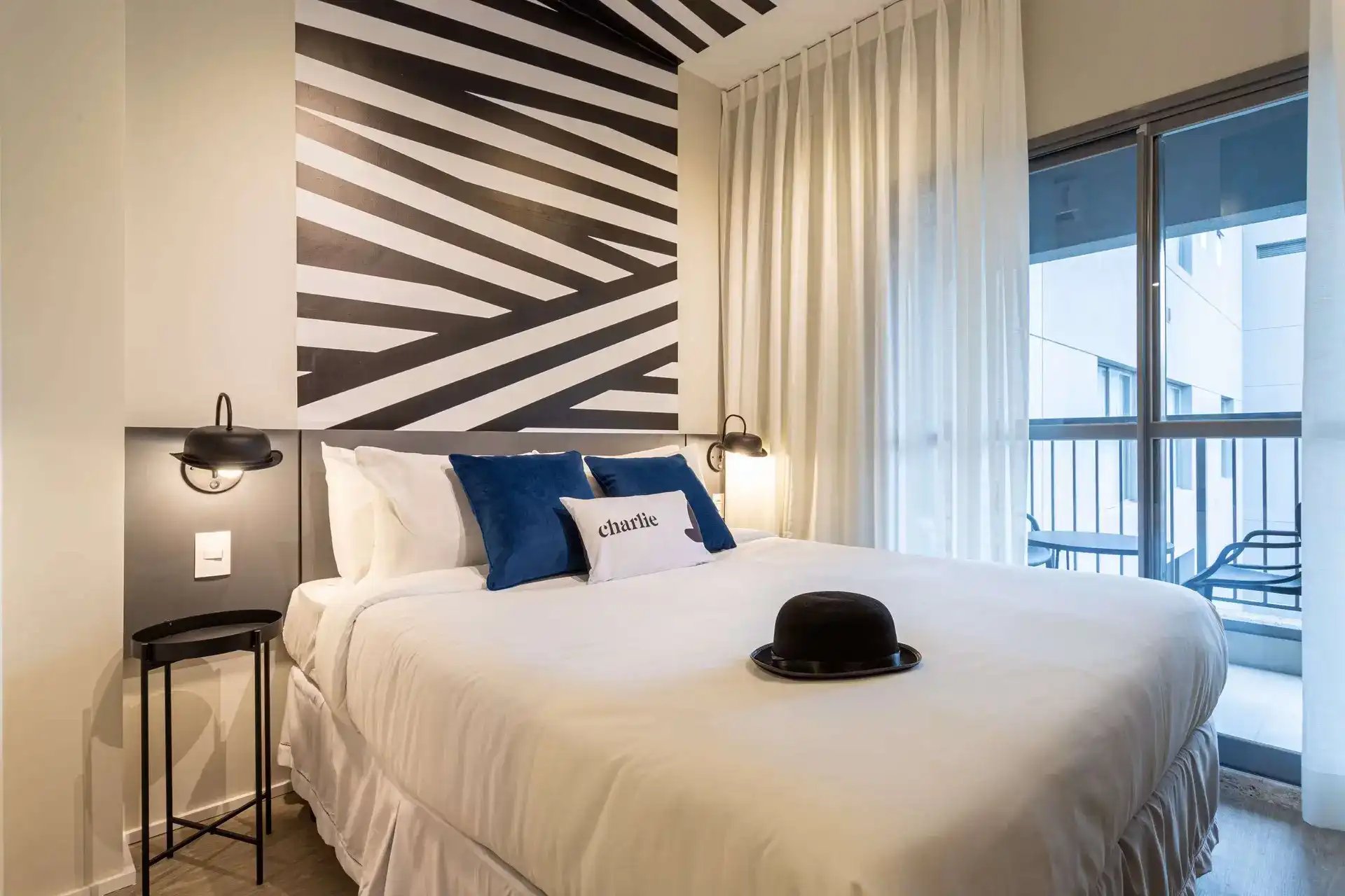 Quarto de hotel moderno com cama confortável, almofadas azuis e decorações elegantes, incluindo uma parede decorativa de listras pretas e brancas e uma varanda com vista.