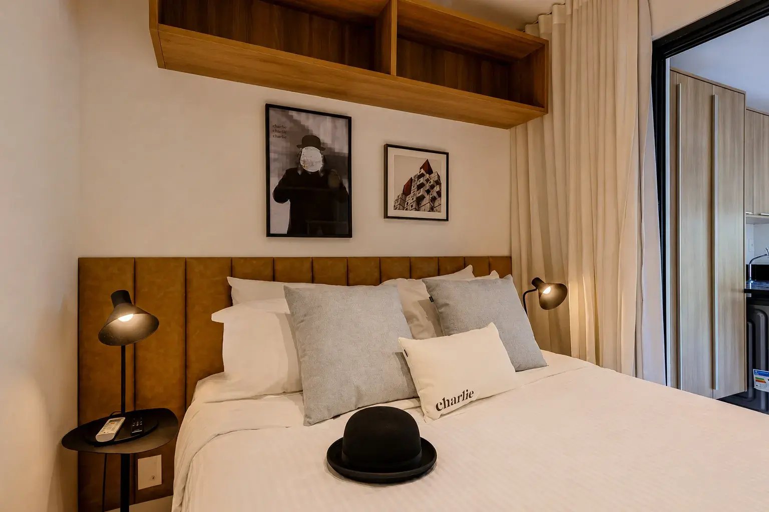 Quarto moderno decorado com cama king ou casal, cabeceira amarela de tecido, almofadas neutras, abajures dourados e chapéu sobre a cama, com janelas e armários ao fundo