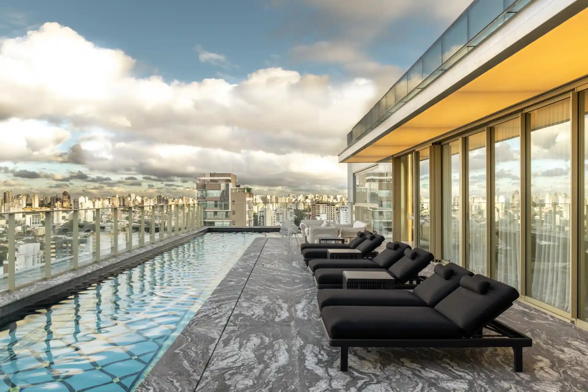 Imagem de uma varanda de cobertura com piscina e vista panorâmica da cidade ao entardecer, com cadeiras de descanso pretas e uma atmosfera de luxo.