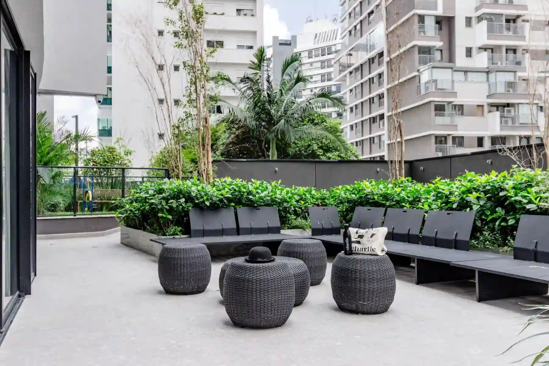 Varanda moderna com móveis pretos de fibra sintética, incluindo cadeiras e pufes, rodeada de vegetação e vista de prédios altos na cidade.
