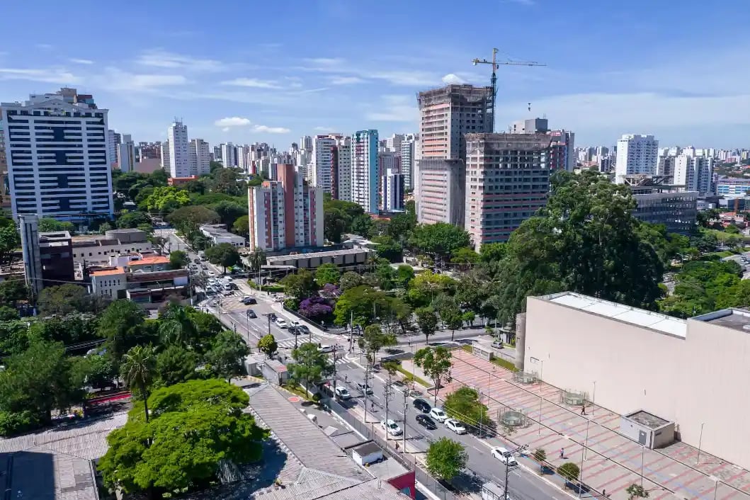 Vista aérea de uma avenida urbana com prédios altos, áreas arborizadas e carros circulando sob céu azul e nuvens, em uma paisagem de cidade em crescimento
