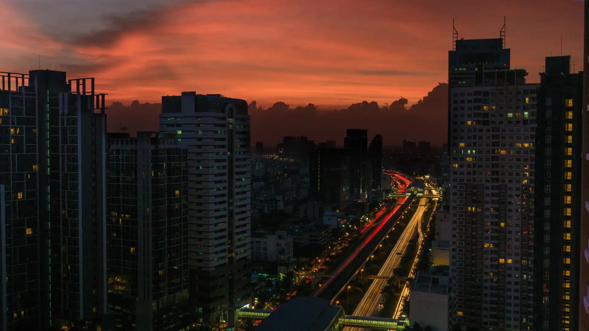 Vista de cidade ao entardecer com prédios altos e céu com cores quentes de pôr do sol, além de luzes de veículos na estrada.