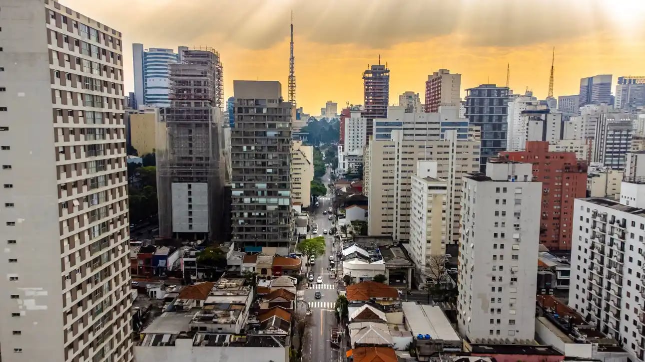 Vista panorâmica de uma avenida em meio a prédios altos e apartamentos, com skyline urbano ao pôr do sol e o céu em tons dourados