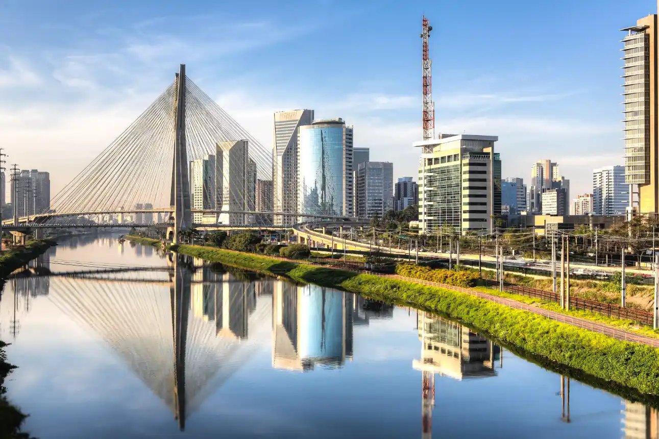 Vista panorâmica de uma cidade moderna com um canal e água refletindo os prédios, incluindo a ponte de cabos com pilar alto, arranha-céus e vegetação à beira do rio ao pôr do sol.